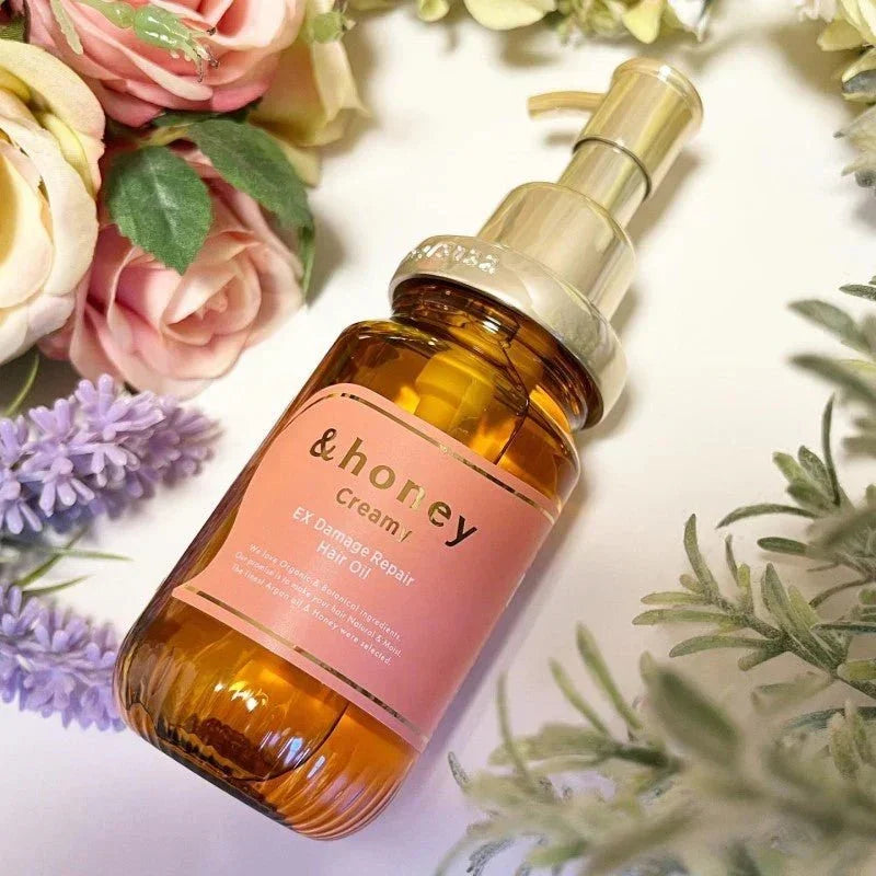 Creamy EX Damage Repair Hair Oil in amber bottle with gold pump, surrounded by pink roses and lavender